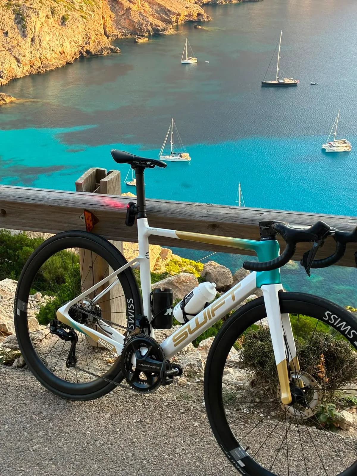 A white and gold Swift resting against a timber rail above a turquoise bay, yachts at anchor below