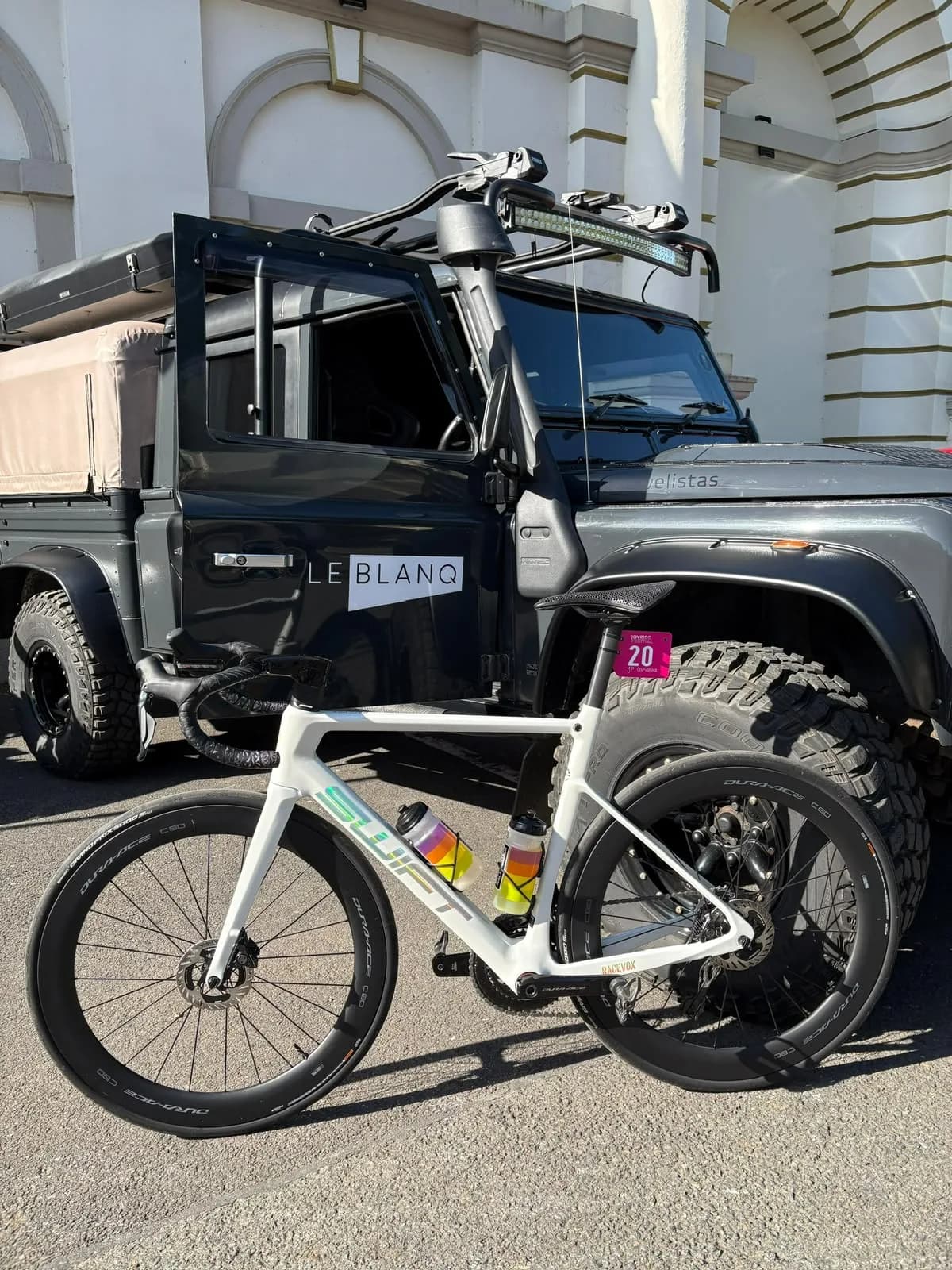 A white Swift on deep-section carbon wheels, leaning against a LeBlanq Land Rover in the morning sun